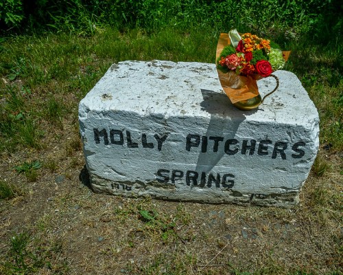 A stone marker reading "Molly Pitcher's Spring" with a bouquet of flowers placed on top, surrounded by grass.