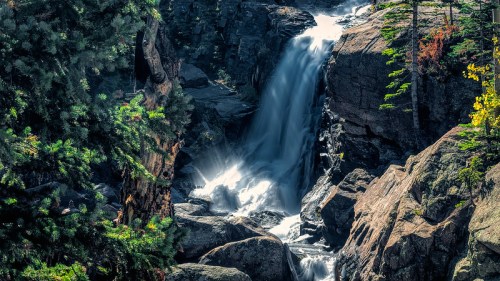 A serene waterfall cascades over rocky terrain, surrounded by lush green trees and sunlight filtering through.