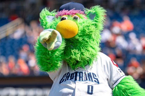 A vibrant green mascot with colorful fur and a baseball cap points playfully at the camera during a game.
