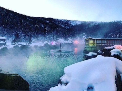 A serene winter scene featuring a steaming hot spring surrounded by snow-covered rocks and trees, with colorful lights.