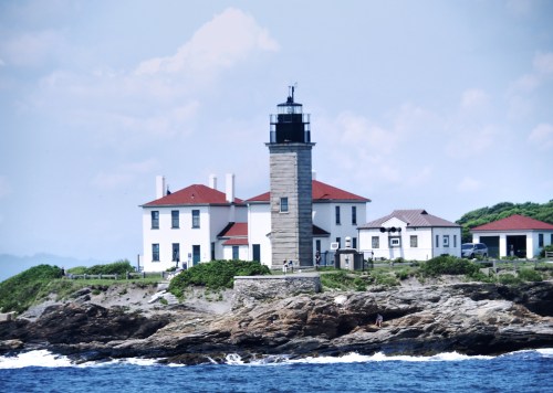 A lighthouse on a rocky shore, surrounded by greenery and a white building with red roofs under a blue sky.