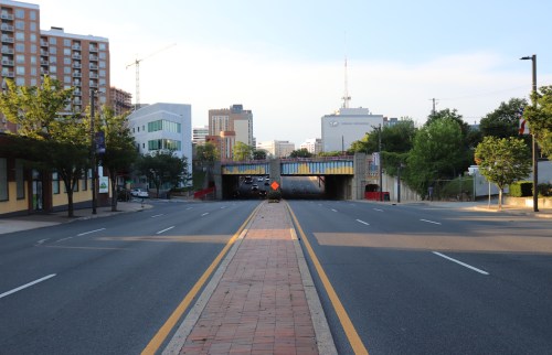 A wide street leads to an underpass, flanked by trees and buildings, with a clear blue sky above.