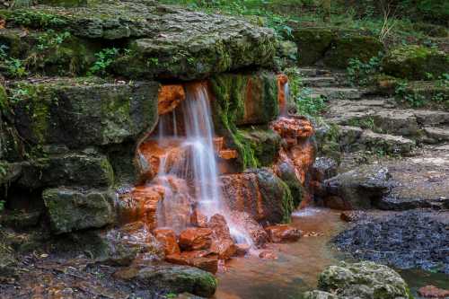 A small waterfall cascades over moss-covered rocks into a tranquil pool, surrounded by lush greenery.