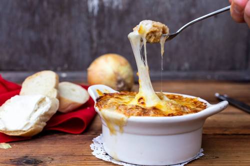 A spoon lifts cheesy onion soup from a white bowl, with bread and an onion in the background on a wooden table.