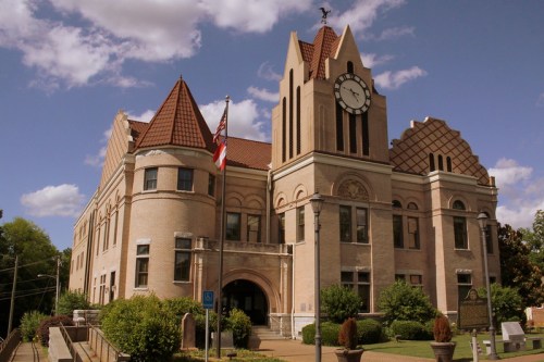 Historic brick building with a clock tower, American flag, and landscaped grounds under a partly cloudy sky.