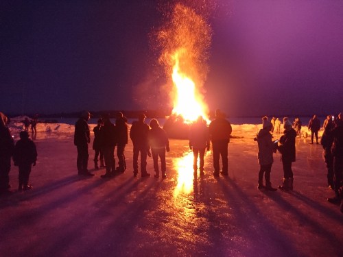A large bonfire illuminates a dark winter night, with silhouettes of people gathered around on an icy surface.