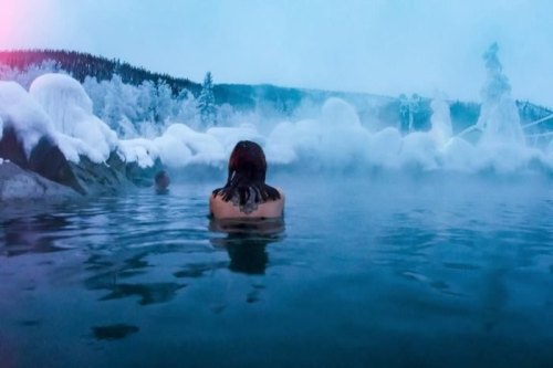 A person relaxes in a steaming hot spring surrounded by snowy landscapes and frosty trees.