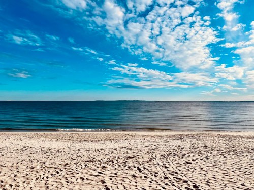 A serene beach scene with soft sand, calm water, and a bright blue sky dotted with clouds.