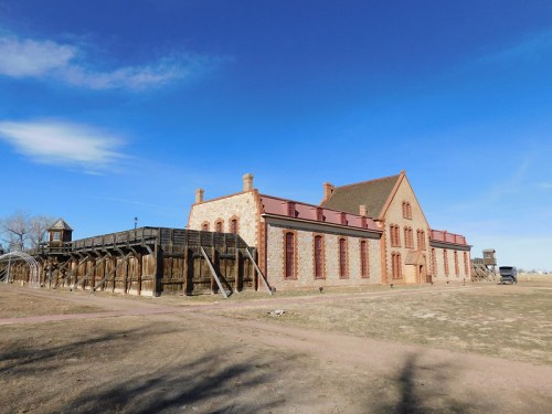 Historic stone building with a red roof, surrounded by open land and a wooden watchtower in the background. Clear blue sky.