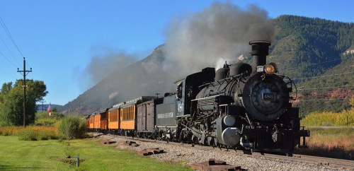 A vintage steam locomotive pulls a train through a scenic landscape with mountains and trees in the background.
