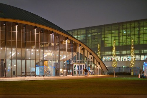 A modern building with a curved glass facade, illuminated at night, surrounded by green grass and festive lights.