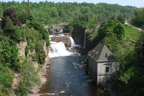 A scenic view of waterfalls cascading into a river, surrounded by lush greenery and a historic building nearby.