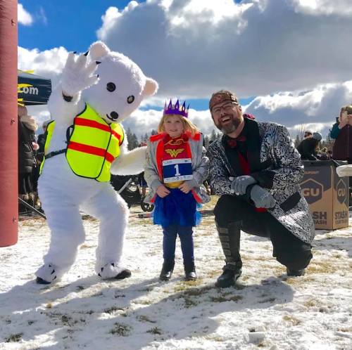 A child in a Wonder Woman costume poses with a bear mascot and a man in a silver coat, all in a snowy outdoor setting.