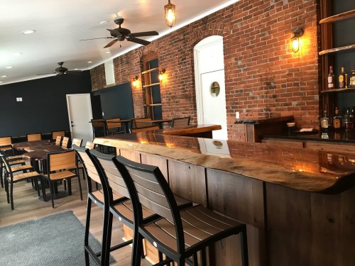 A modern bar interior with wooden seating, a polished counter, and exposed brick walls, featuring warm lighting.