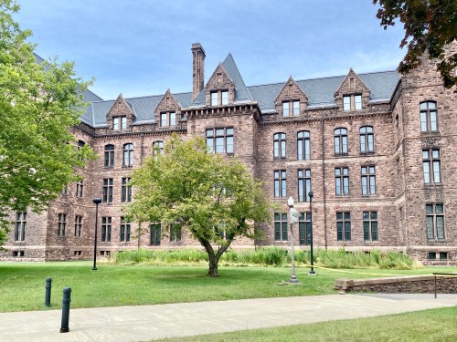 Historic stone building with multiple windows, surrounded by green grass and trees under a cloudy sky.