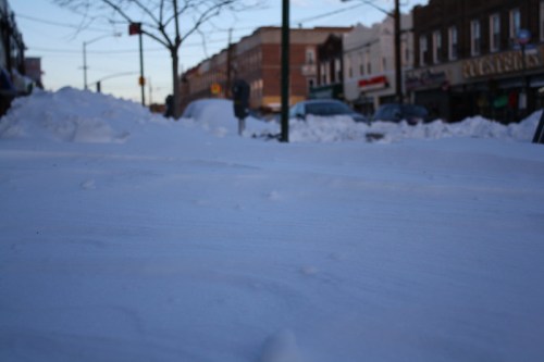 A snowy street scene with deep snow drifts and buildings in the background, captured in soft morning light.