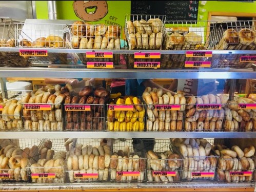 A display case filled with various bagels in baskets, labeled with flavors like blueberry, garlic, and chocolate chip.