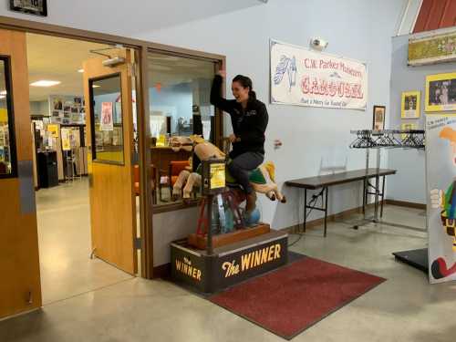 A person playfully rides a vintage mechanical horse inside a museum, with colorful displays in the background.