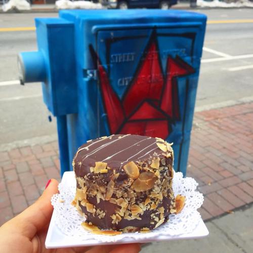 A hand holding a chocolate-covered dessert with nuts, in front of a blue utility box featuring red graffiti.