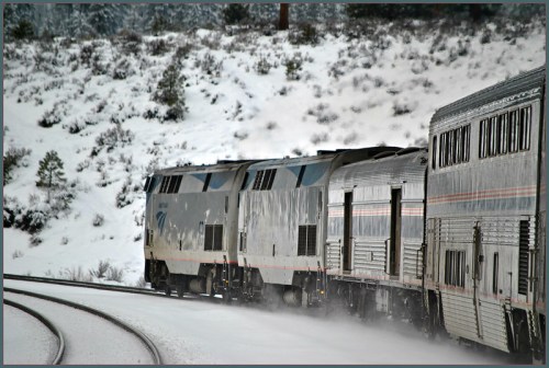 A train travels along snowy tracks, surrounded by a winter landscape of trees and hills.