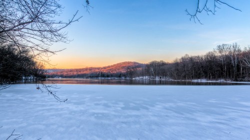 A serene winter landscape featuring a frozen lake, snow-covered trees, and a sunset over distant hills.