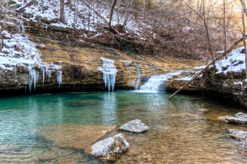 A serene winter scene featuring a clear blue pool, icy waterfalls, and snow-covered rocks in a wooded area.