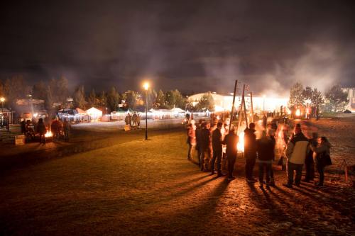 A nighttime scene with people gathered around bonfires, tents in the background, and smoke rising in the air.