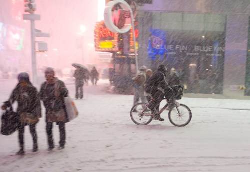 A snowy scene in Times Square with pedestrians and a cyclist navigating through heavy snowfall.