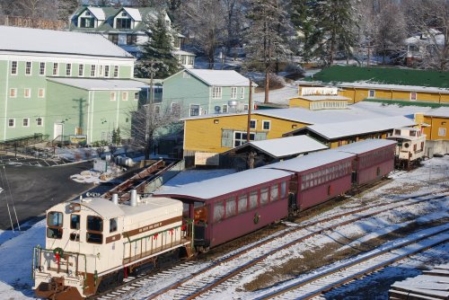 A vintage train with maroon cars parked near colorful buildings, surrounded by snow-covered ground and trees.