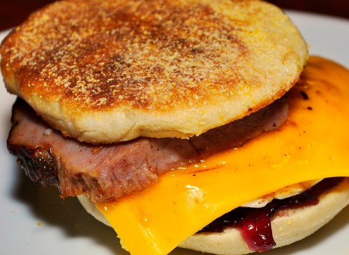 A toasted English muffin sandwich with sliced brisket, melted cheddar cheese, and a layer of jam.