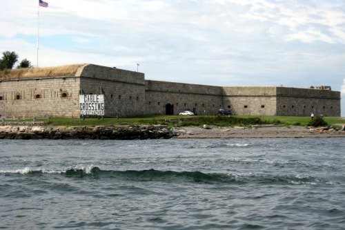 Historic stone fort with a grassy area, near water, featuring a large sign that reads "Whale Crossing."
