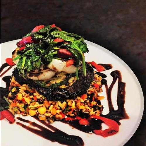 A gourmet dish featuring grilled vegetables, a portobello mushroom, topped with greens and drizzled with sauce on a plate.