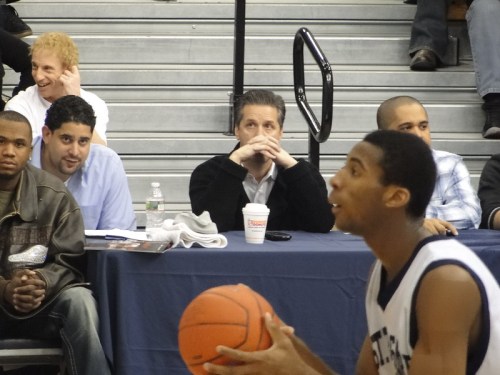 A basketball player prepares to shoot while spectators, including a focused man, watch from the sidelines.