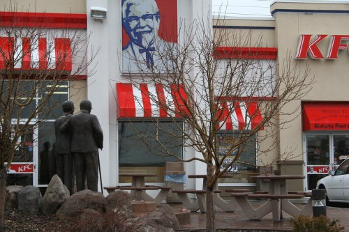 A KFC restaurant with a Colonel Sanders mural, featuring a statue of two people in front and bare trees nearby.