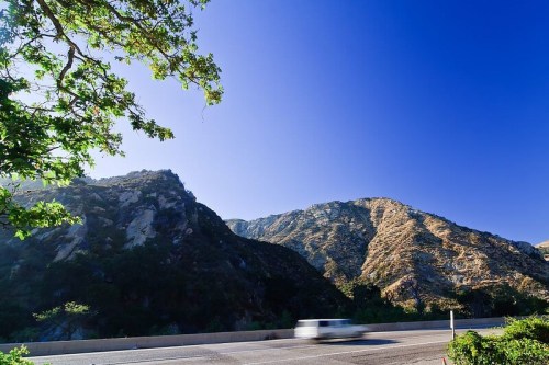 A scenic view of mountains under a clear blue sky, with a blurred white vehicle passing on the road.