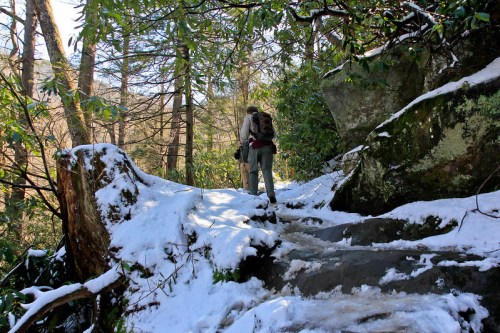 A person hiking on a snowy trail surrounded by trees and rocky terrain.