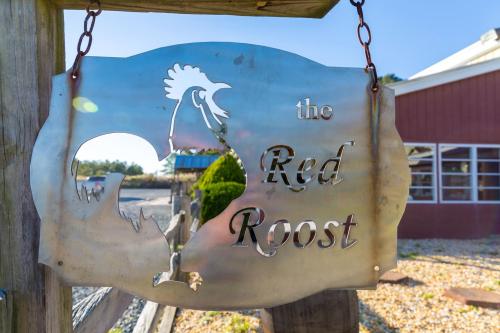 Metal sign reading "the Red Roost" with a rooster silhouette, hanging near a rustic building on a sunny day.