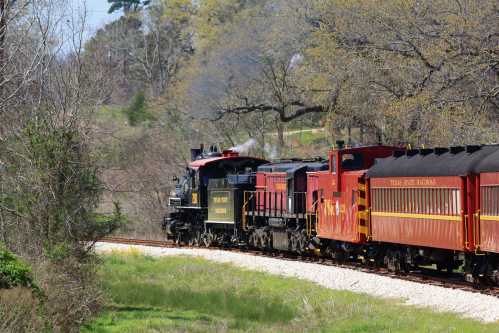 A vintage steam locomotive pulls a red train along a scenic, tree-lined track on a sunny day.