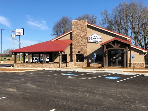 Exterior of Hoskins BBQ restaurant featuring a stone facade, red roof, and parking lot on a sunny day.