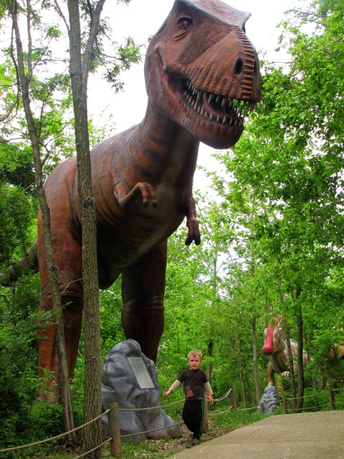 A child walks along a path beside a large dinosaur statue in a green, wooded area.