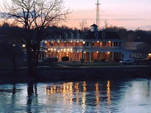 A riverside building at sunset, reflecting in the water, with trees and a communication tower in the background.