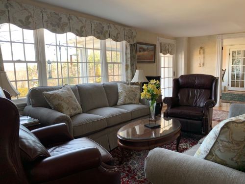 Cozy living room with large windows, floral curtains, a coffee table, and comfortable seating.