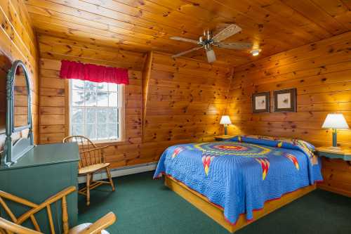 Cozy wooden bedroom with a blue quilted bedspread, rocking chair, and two lamps beside the bed. Natural light from the window.