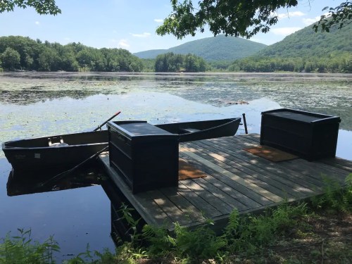 A serene lake scene with a wooden dock, two boats, and lush green mountains in the background under a clear blue sky.