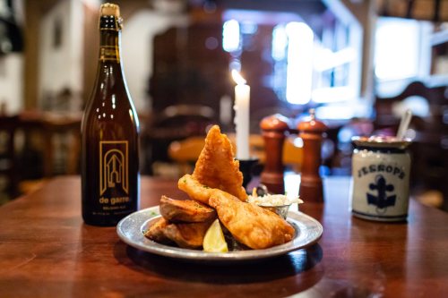 A plate of fried fish and chips with lemon, a candle, and a bottle of de garre beer on a wooden table.