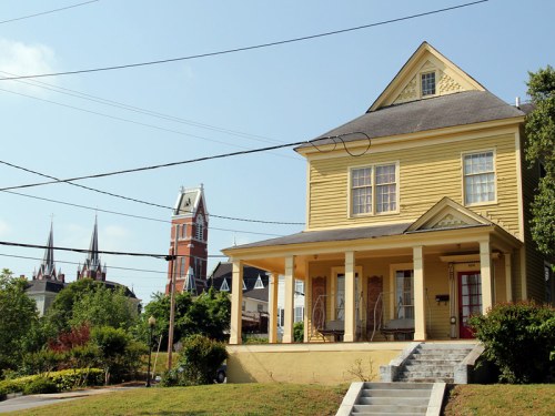 A yellow house with a porch, surrounded by greenery, with church steeples visible in the background under a clear blue sky.