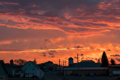 A vibrant sunset paints the sky in orange and purple hues over a small town with buildings and a church steeple.