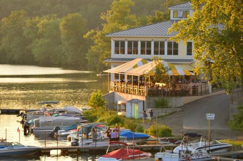 A lakeside restaurant with a striped awning, surrounded by boats and trees, during a golden sunset.