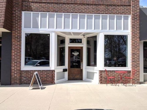 A brick storefront with large windows, featuring a wooden door and a small outdoor seating area with red chairs.
