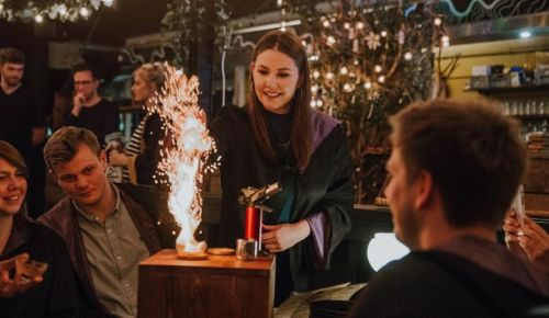 A woman in a dark robe ignites a sparkler at a lively gathering, with friends watching in a cozy, festive setting.